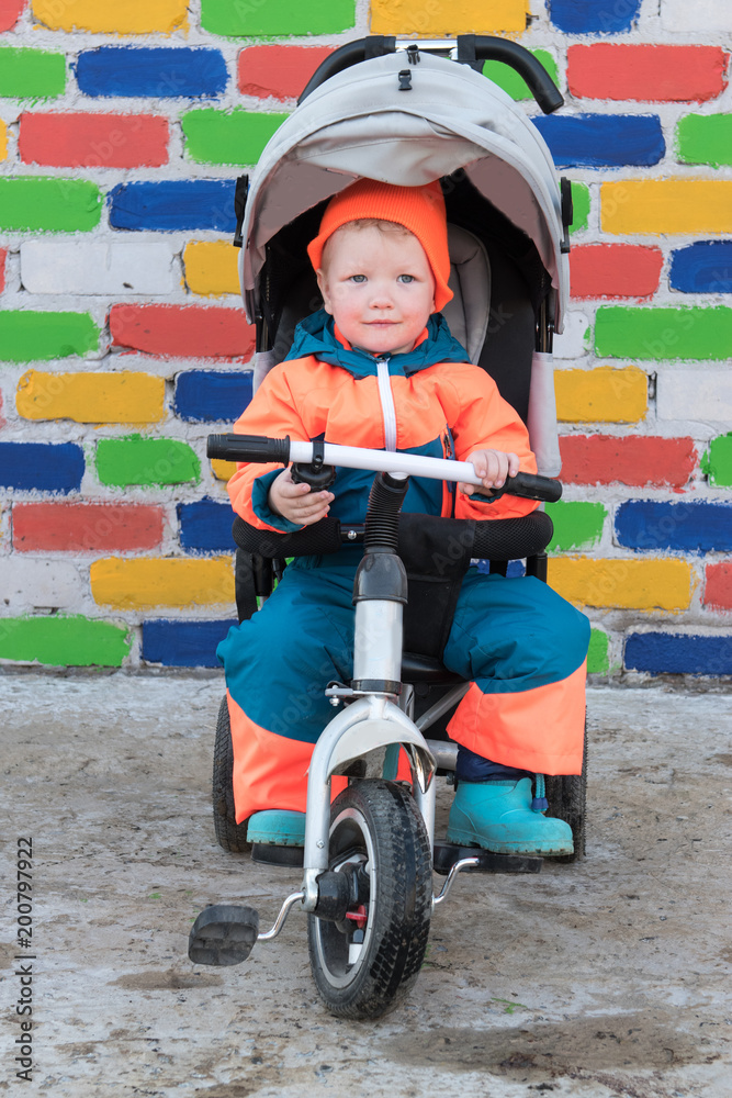 Happy boy in bright suit is riding children's bicycle wheelchair against background of multicolored brick wall. Romantic lighting with sunlight reflections. Lens flare effect without post prodaction Happy boy in bright suit is riding children's bicycle wheelchair against background of multicolored brick wall. Romantic lighting with sunlight reflections. Lens flare effect without post prodaction