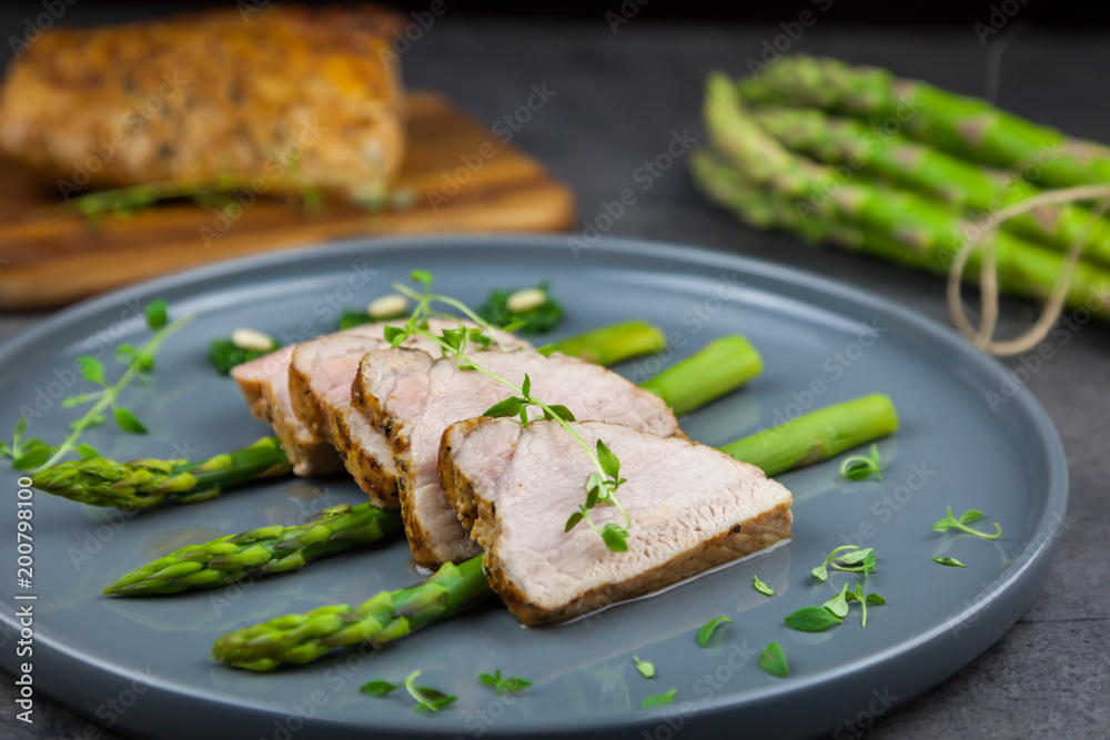 Slices of roasted pork tenderloin on asparagus with pesto from kale and thyme on the grey plate. Asparagus and all pork tenderloin on the background
