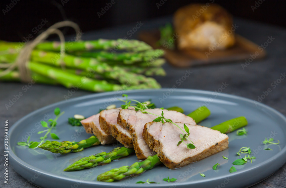 Slices of roasted pork tenderloin on asparagus with pesto from kale and thyme on the grey plate. Asparagus and all pork tenderloin on the background