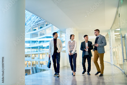 Group of young white collar workers walking along corridor of modern office building and having small talk