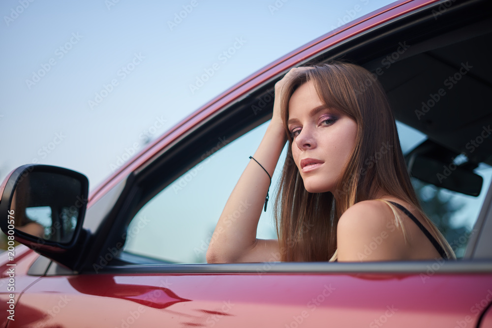 Concept: strong independent young woman in car. Beautiful serious girl sit behind the wheel at twilight looking through the window.