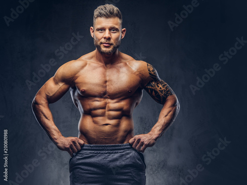 A handsome shirtless tattooed bodybuilder with stylish haircut and beard, wearing sports shorts, posing in a studio. Isolated on a dark background
