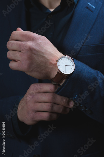 A dark portrait of a man dressed in an expensive suit, adjusting an elegant wrist watch