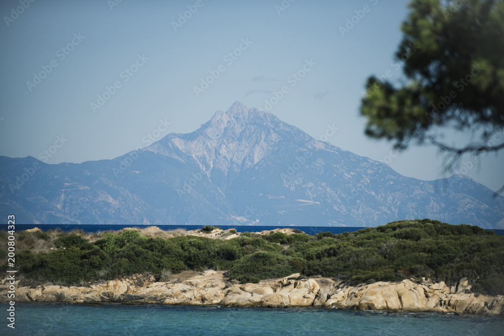 Beautiful landscape with high mountain, covered with snow. Light rocky top of mountain and blue sky, horizon, skyline. Sea and stony seashore landshaft covered with green plants. Nature on sunny day.