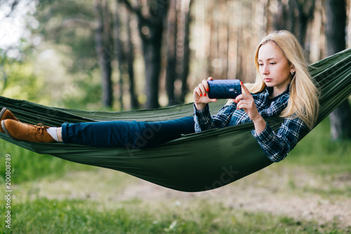 Young beautiful blonde girl in hammock and making selfie. Pretty woman leisure lifestyle portrait at nature ountdoor. Female relax in forest. Adorable teen face expression. Tourism in summer camp. photo