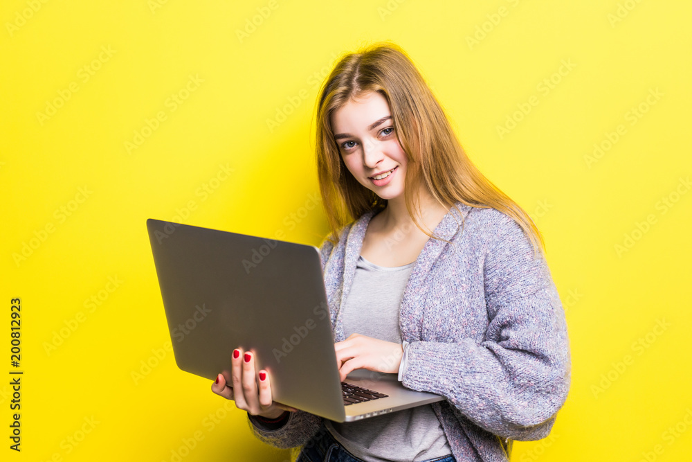 Portrait of a smiling teenage girl holding laptop computer isolated on a yellow background and looking at camera Portrait of a smiling teenage girl holding laptop computer isolated on a yellow background and looking at camera