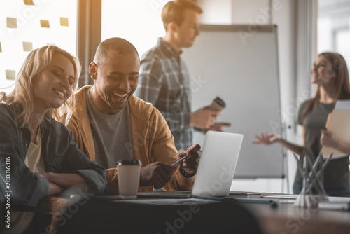 Man and woman watching interesting video at computer during coffee pause. Colleagues communicating on background
