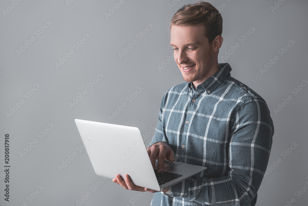Waist up portrait of handsome man standing with laptop in hands and looking at screen with joy. Isolated on background