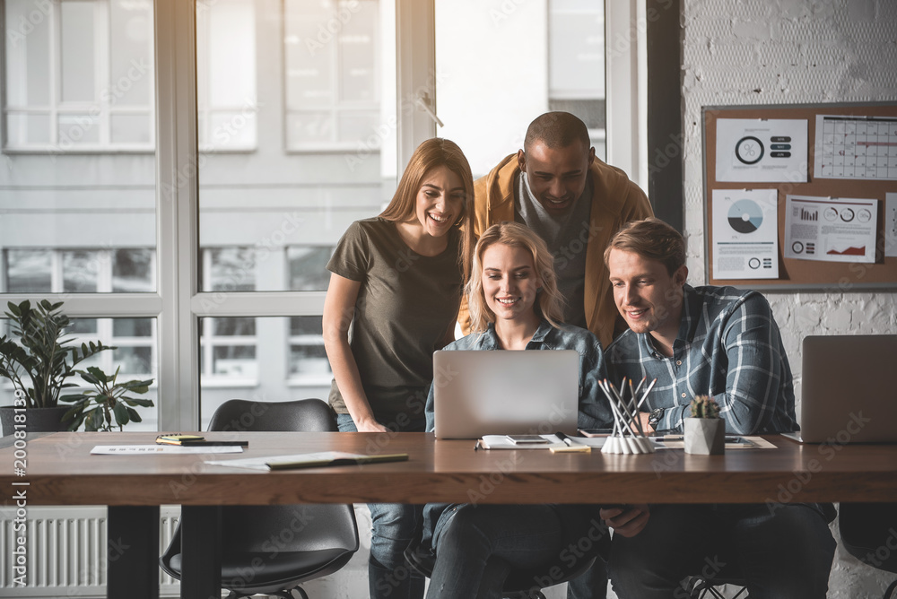 Four young people working in office. They are looking at notebook screen with pleasure. Copy space in left side Four young people working in office. They are looking at notebook screen with pleasure. Copy space in left side