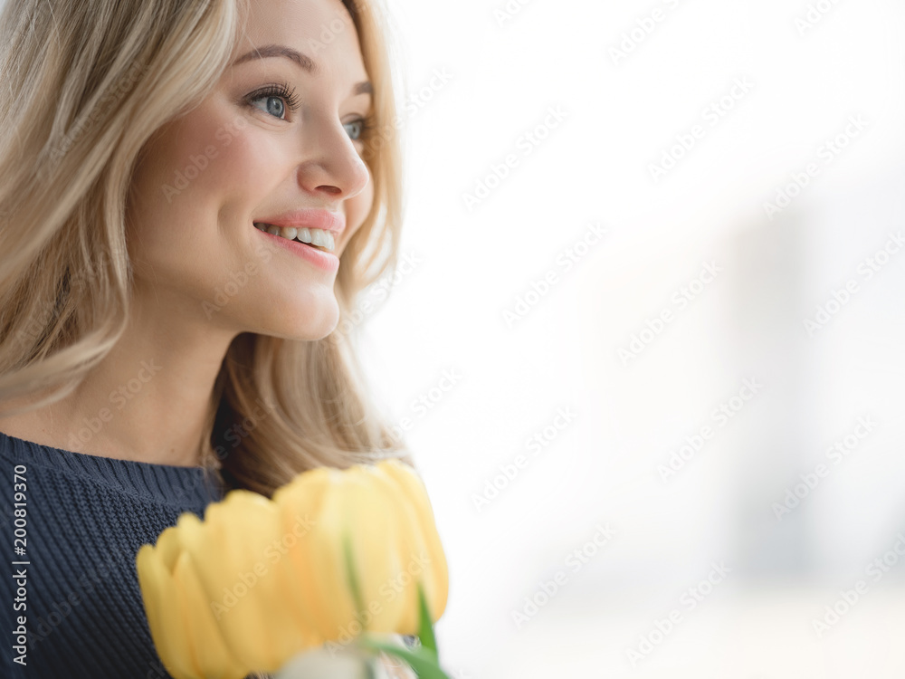 Positive young woman holding yellow tulips and smiling. She is standing near the window with relaxation. Copy space Positive young woman holding yellow tulips and smiling. She is standing near the window with relaxation. Copy space