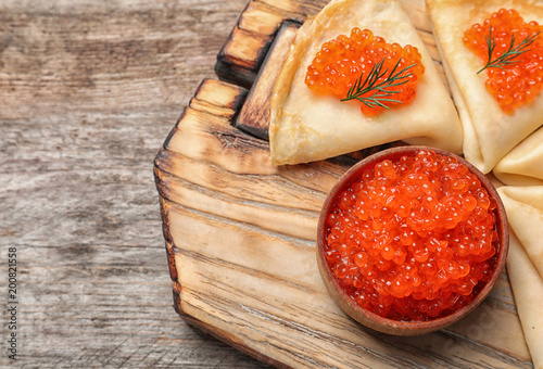 Bowl with delicious red caviar and thin pancakes on wooden board