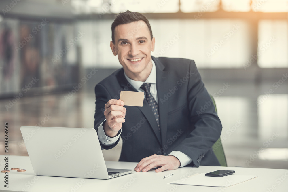 Portrait of beaming businessman demonstrating visiting card while working with laptop at desk. Happy worker during occupation concept Portrait of beaming businessman demonstrating visiting card while working with laptop at desk. Happy worker during occupation concept