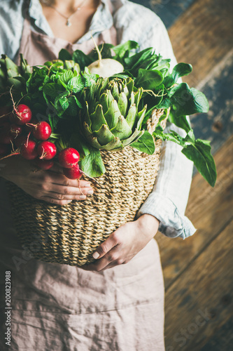 Female farmer wearing pastel linen apron and shirt holding basket with fresh seasonal vegetables in her hands, selective focus. Organic produce or local market concept