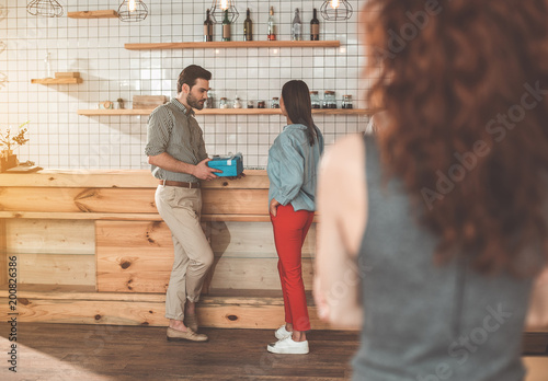 Seductive man giving present to young girl in coffee shop. Jealous woman is looking at them from the side