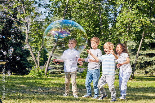 Multi-ethnic group of children having fun with big soap bubbles while spending warm summer day at public park illuminated with sunbeams
