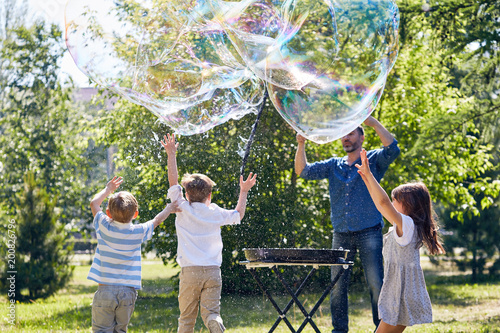 Bearded middle-aged animator making big soap bubbles while cheerful little children trying to catch them, green park illuminated with sunbeams on background photo