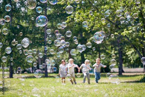 Green public park illuminated with sunbeams, colorful soap bubbles flying in air, group of little children playing active games