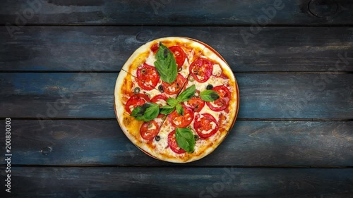 Perfect pizza margarita with tomato slices, olives and basil leaves rotating on a dark wooden background. Top view