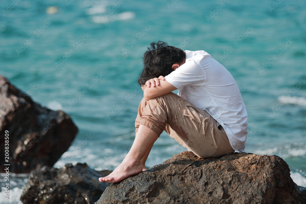Sorrowful stressed Asian man sit and hug knees up to the chest at the rock of seashore.