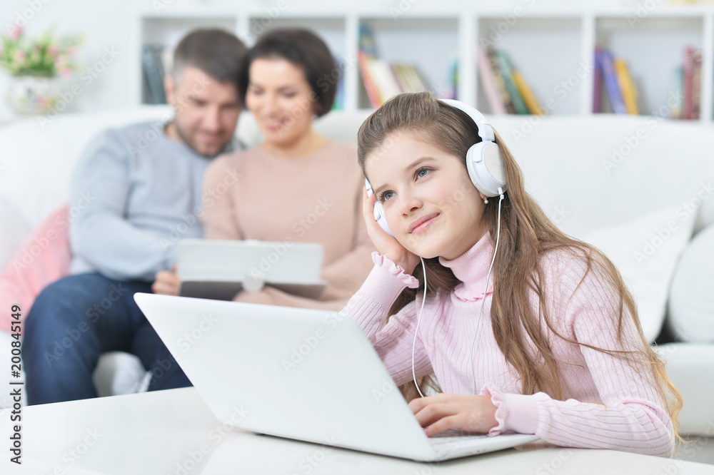 Happy girl sitting at table with laptop with her parents on bac Happy girl sitting at table with laptop with her parents on bac