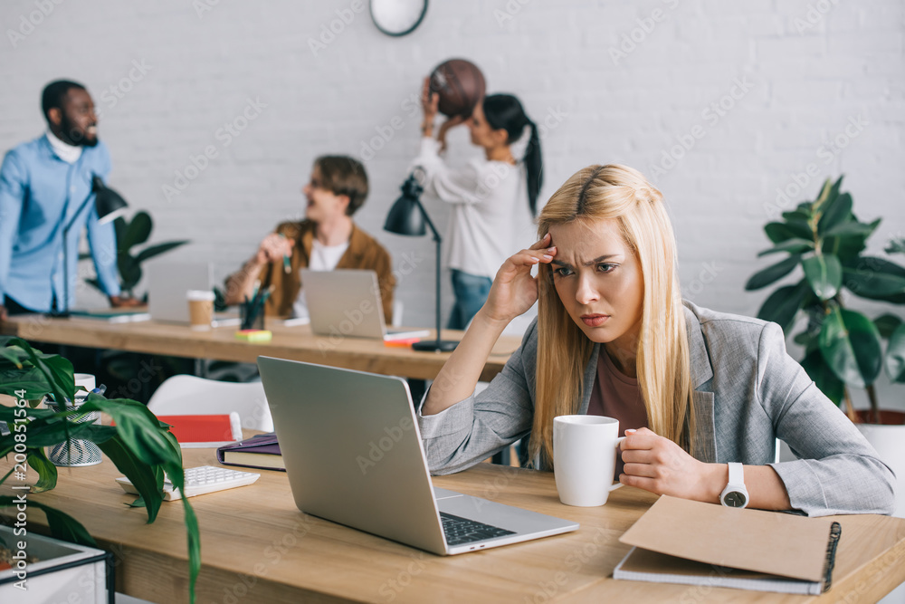 Fototapeta premium frustrated businesswoman with coffee mug watching on laptop screen and colleagues with ball behind