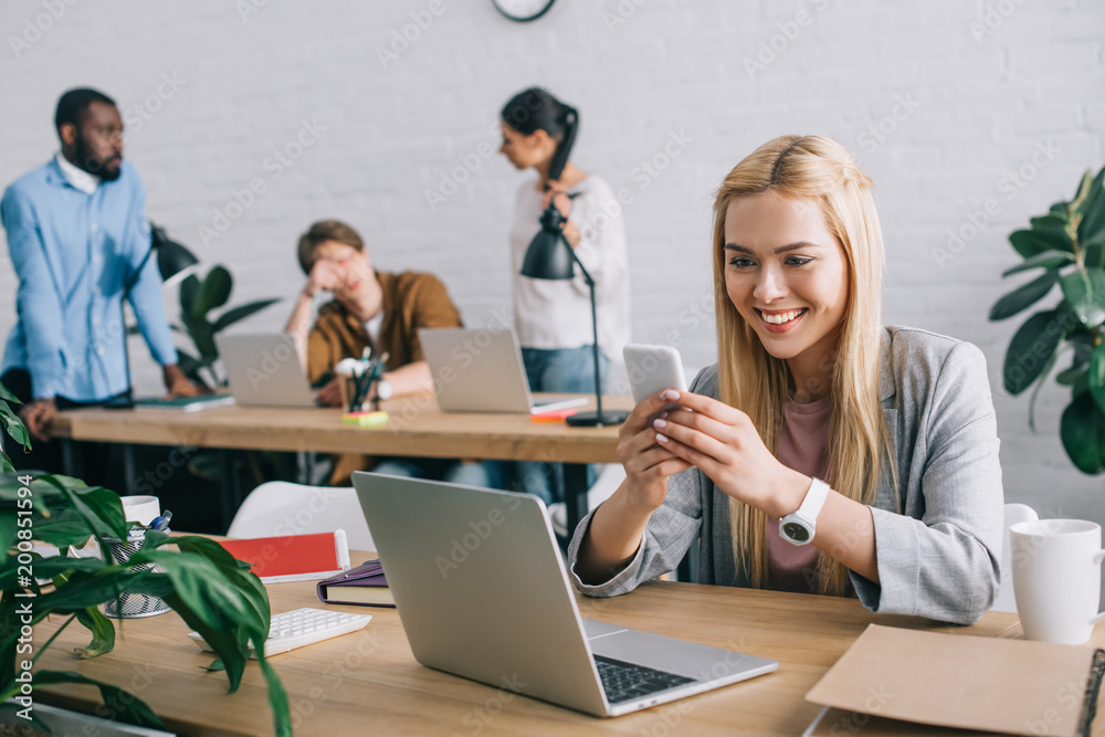 Fototapeta premium happy businesswoman typing on smartphone and colleagues standing behind in modern office