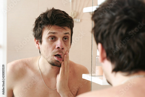 Tired man who has just woken up looks at his reflection in the mirror slaps his cheek with his hand.