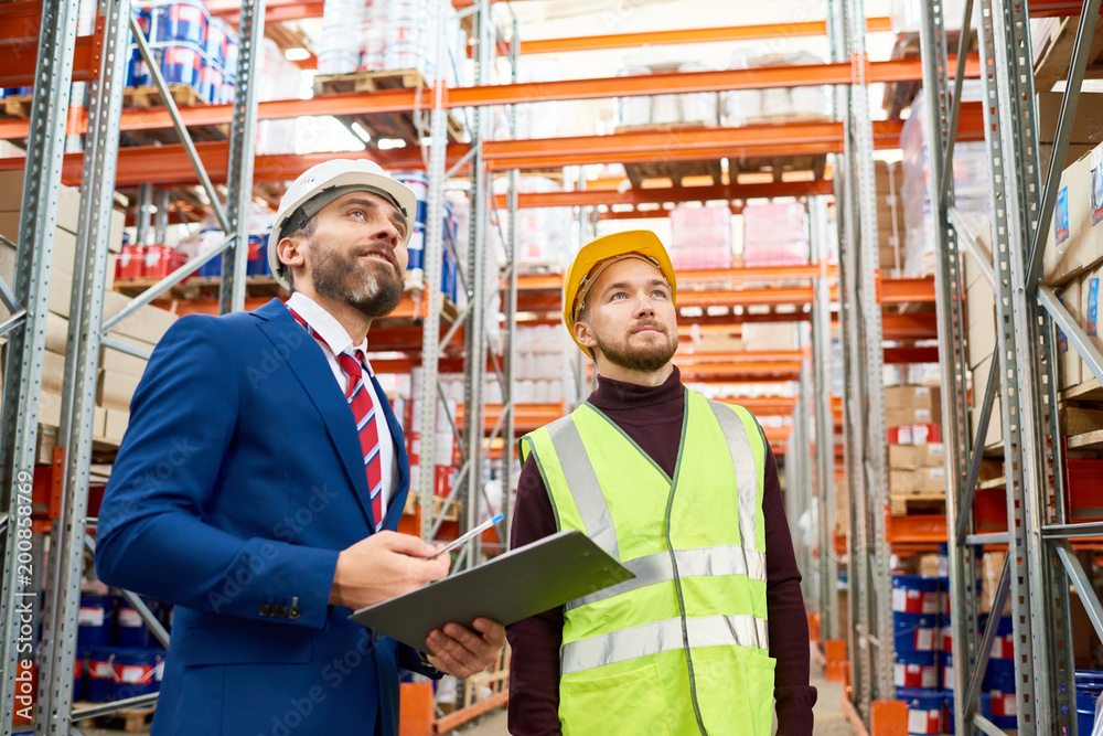 Waist up portrait of warehouse manager holding clipboard talking to worker wearing hardhat and reflective jacket while discussing stock inventory