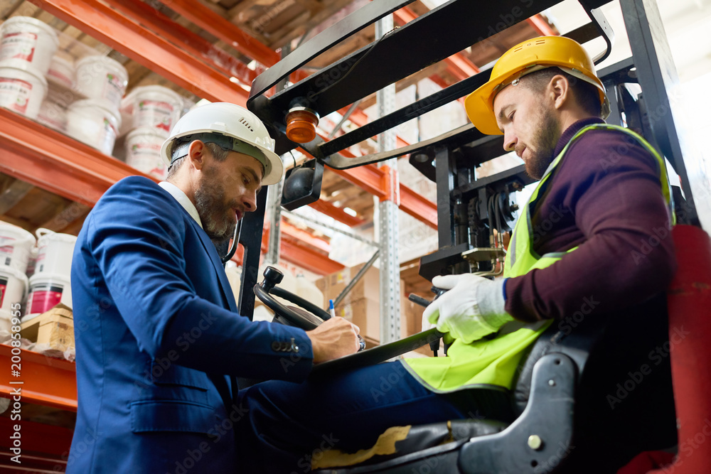 Side view portrait of mature businessman talking to warehouse worker sitting in forklift car Side view portrait of mature businessman talking to warehouse worker sitting in forklift car