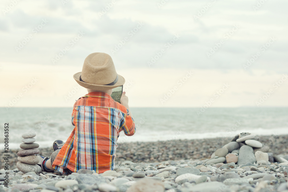 Little boy takes pictures on the smart phone. Sits on a pebble beach