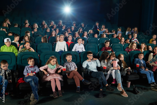 Frontview of people sitting in the cinema hall and watching movie. Boys and girls watching interesting movie and looking very emotional  frightened and exited. Children wear colorful trendy clothes.