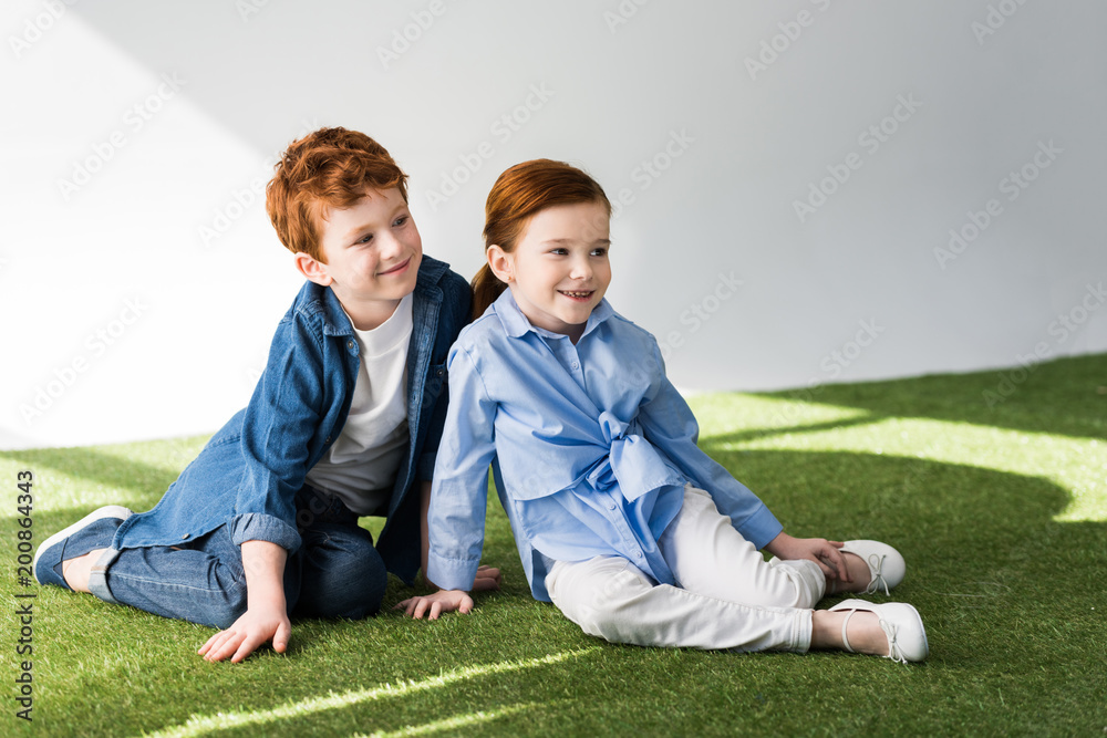 adorable happy redhead kids sitting together on grass and looking away on grey