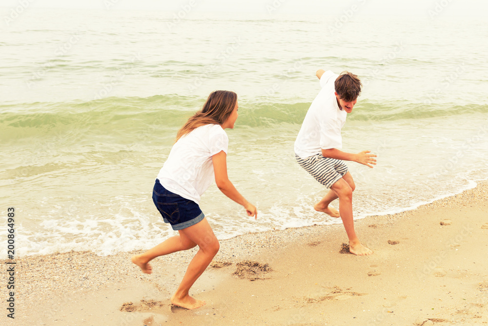 Two teenagers: a girl and a boy with blond hair, dressed in white T-shirts they run, laugh and play in the sea beach. Two teenagers: a girl and a boy with blond hair, dressed in white T-shirts they run, laugh and play in the sea beach.