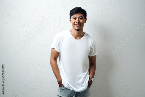 Smiling asian guy in white blank t-shirt, grunge wall, studio portrait