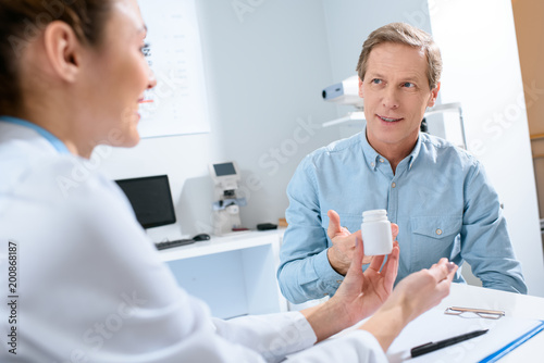 female doctor holding bottle of pills and talking with mature patient in clinic
