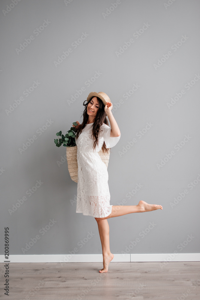 beautiful young brunette girl in white short dress, wearing hat, with summer bag in hand on gray background. Let's go shopping beautiful young brunette girl in white short dress, wearing hat, with summer bag in hand on gray background. Let's go shopping