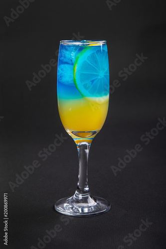 A multi-colored, two-layered yellow and blue cocktail in a high glass with ice cubes with the taste of pineapple, orange and a slice of lemon. Side view. Isolated black background. Drink for the menu