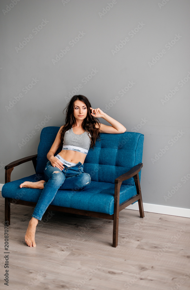 beautiful young brunette girl sitting on blue couch in blue jeans on gray background. Sports girl is resting beautiful young brunette girl sitting on blue couch in blue jeans on gray background. Sports girl is resting