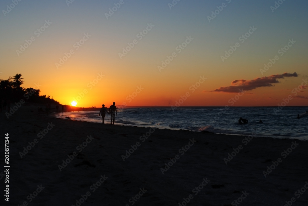 People are holding hands and walking on the beach during sunset. Sunset on the beach in Cuba. Orange sun on the horizon. People are holding hands and walking on the beach during sunset. Sunset on the beach in Cuba. Orange sun on the horizon.