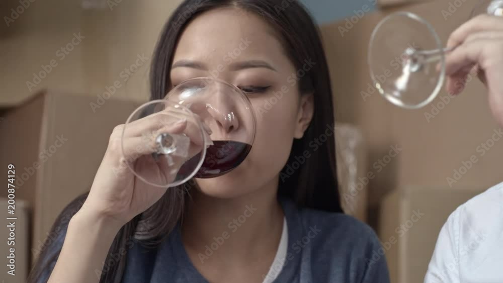 Medium shot of happy Asian couple sitting on floor with glasses of wine and toasting, then kissing and drinking; pile of cardboard boxes in background Medium shot of happy Asian couple sitting on floor with glasses of wine and toasting, then kissing and drinking; pile of cardboard boxes in background