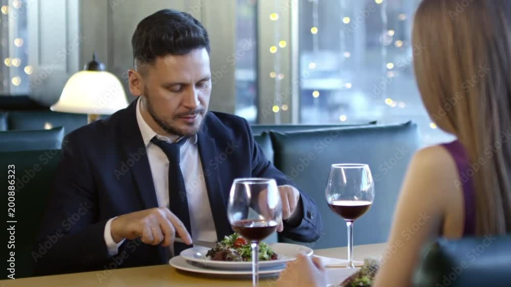 PAN of cheerful bearded man in suit and necktie sitting at table and eating salad while chatting with unrecognizable woman on date in restaurant PAN of cheerful bearded man in suit and necktie sitting at table and eating salad while chatting with unrecognizable woman on date in restaurant