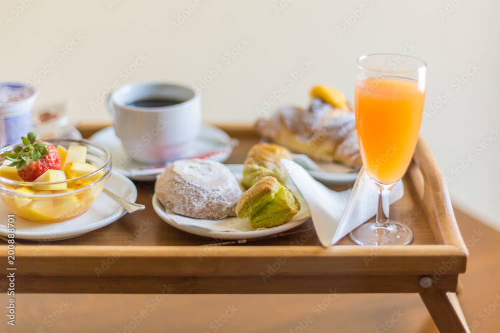 Breakfast on a wooden tray. Green puff pastry, bowl with fruit, bun with icing sugar, coffee cup, croissant, glass of orange juice