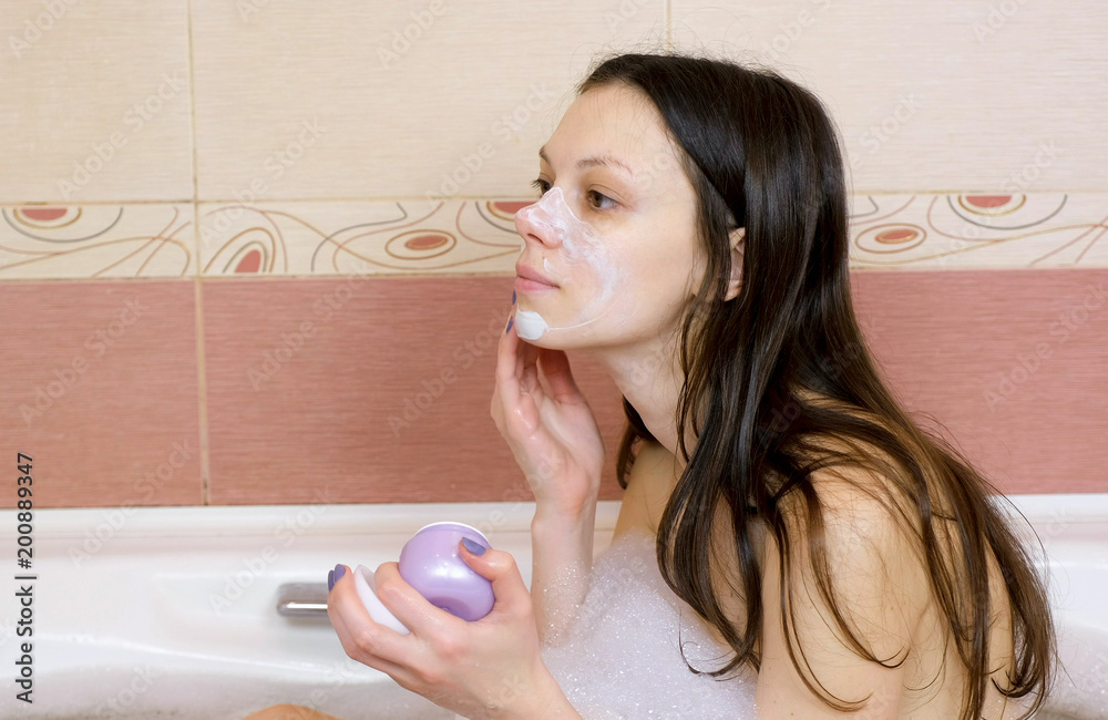 Brunette woman puts on a face mask of blue clay sitting in the bathroom. Face close-up.