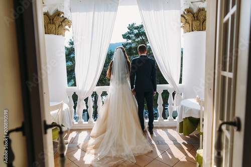 Newlyweds standing on white balcony and holding hands, rear view. The bride in a beautiful white dress with long hair and a veil embraces the groom in a black classic suit.