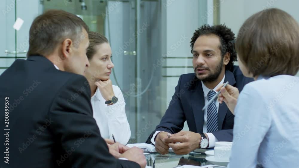 PAN shot of diverse team of businessmen and businesswomen sitting at table in modern office center and discussing ideas PAN shot of diverse team of businessmen and businesswomen sitting at table in modern office center and discussing ideas