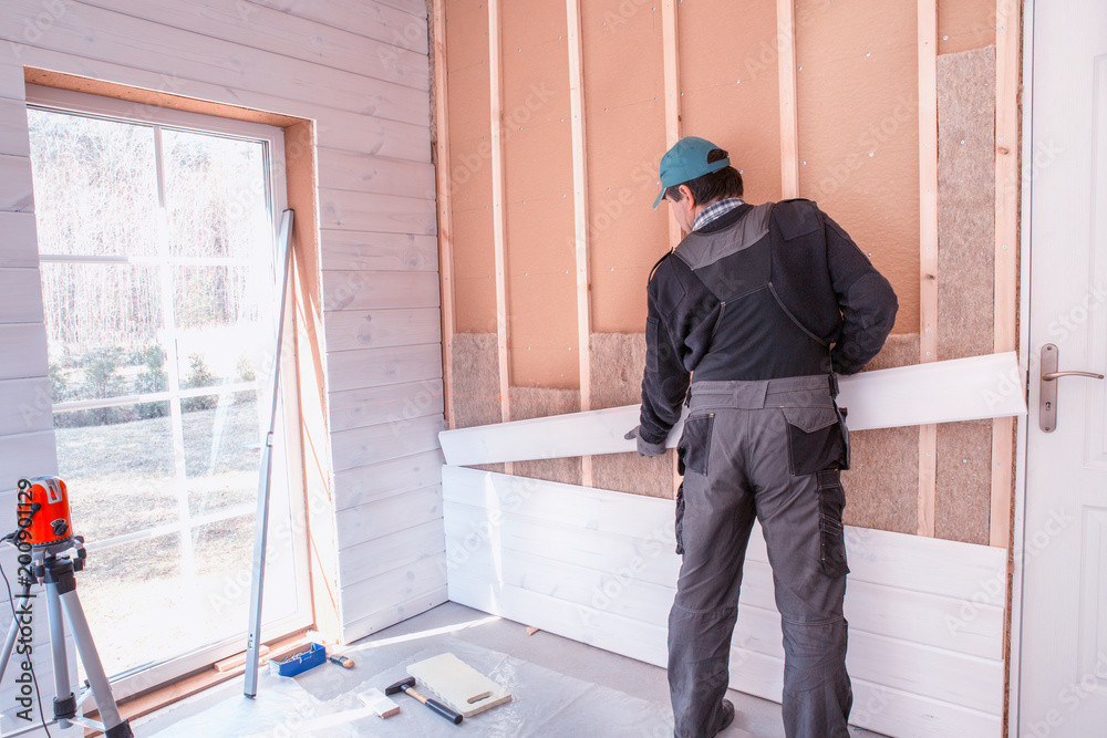 The worker makes finishing works of walls with a white wooden board, using laser line level. Building heat-insulating eco-wooden frame house with wood fiber plates. Latvia
