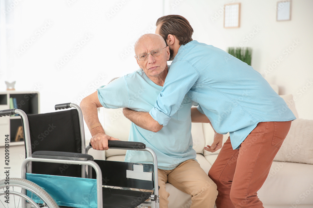 Young caregiver helping senior man to sit in wheelchair at home Young caregiver helping senior man to sit in wheelchair at home