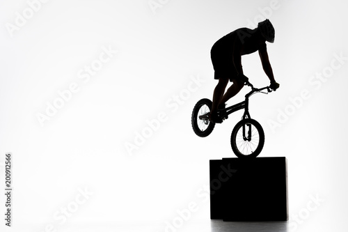 silhouette of trial cyclist performing stunt while balancing on cube on white photo