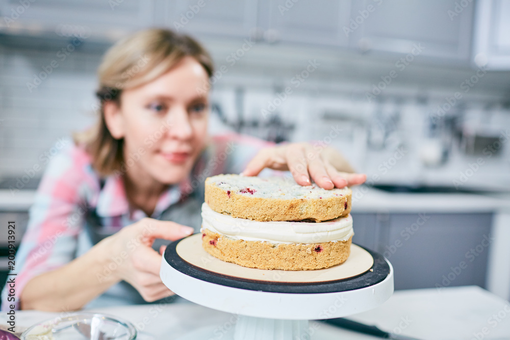 Defocused Caucasian female cook finishing appetizing layer cake with cream in kitchen