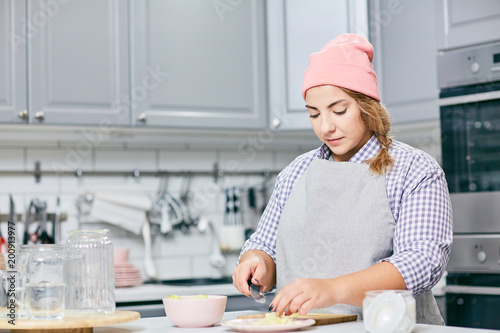 Young concentrated Caucasian woman in apron and hat cutting pear in modern kitchen